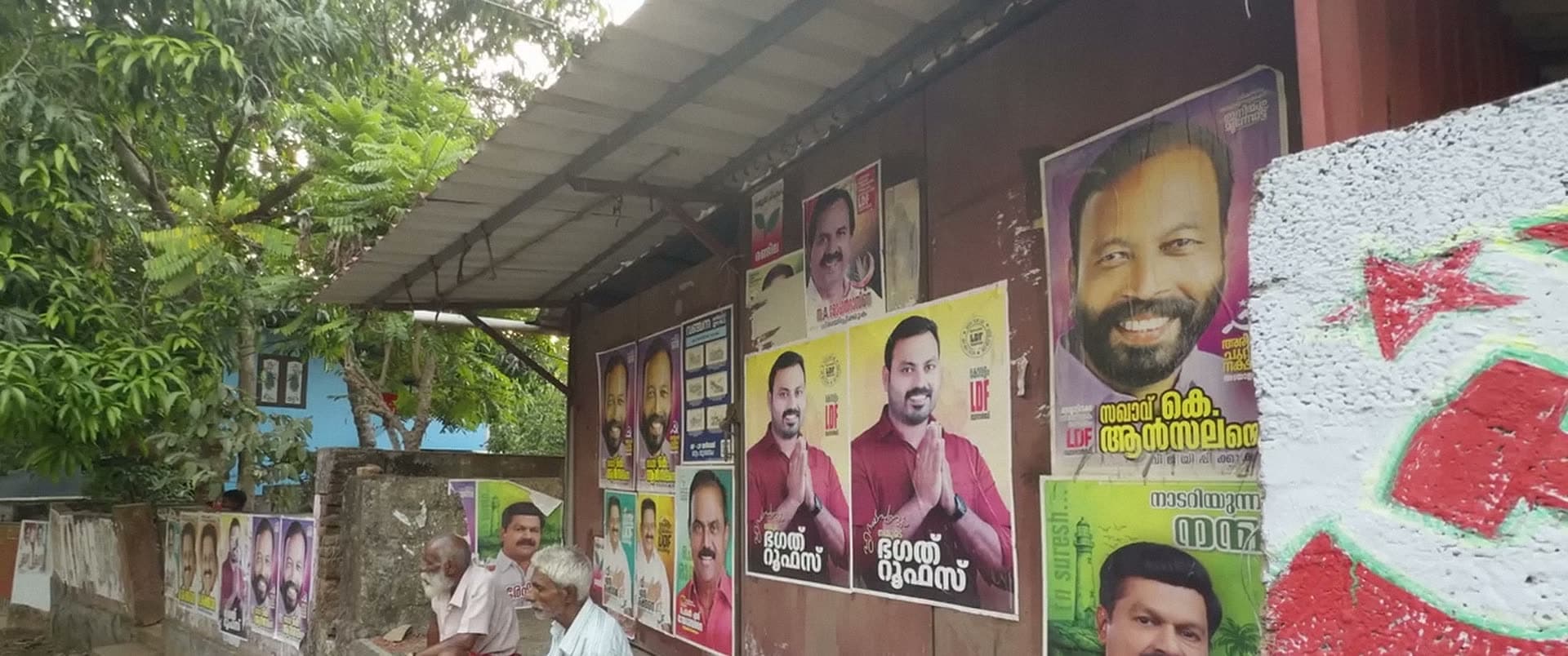 Men sit in the shade of a tin-roofed shelter whose walls are covered in posters, a red star painted beside them.