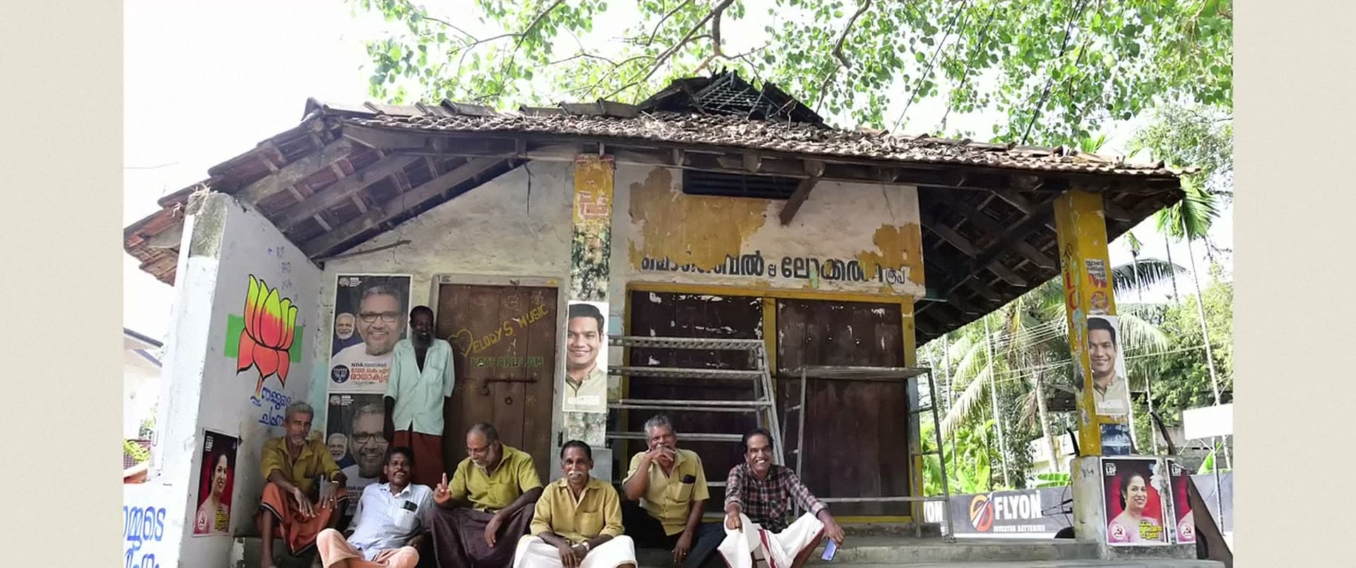A group of men sit on the veranda of a small tiled building pasted with posters.