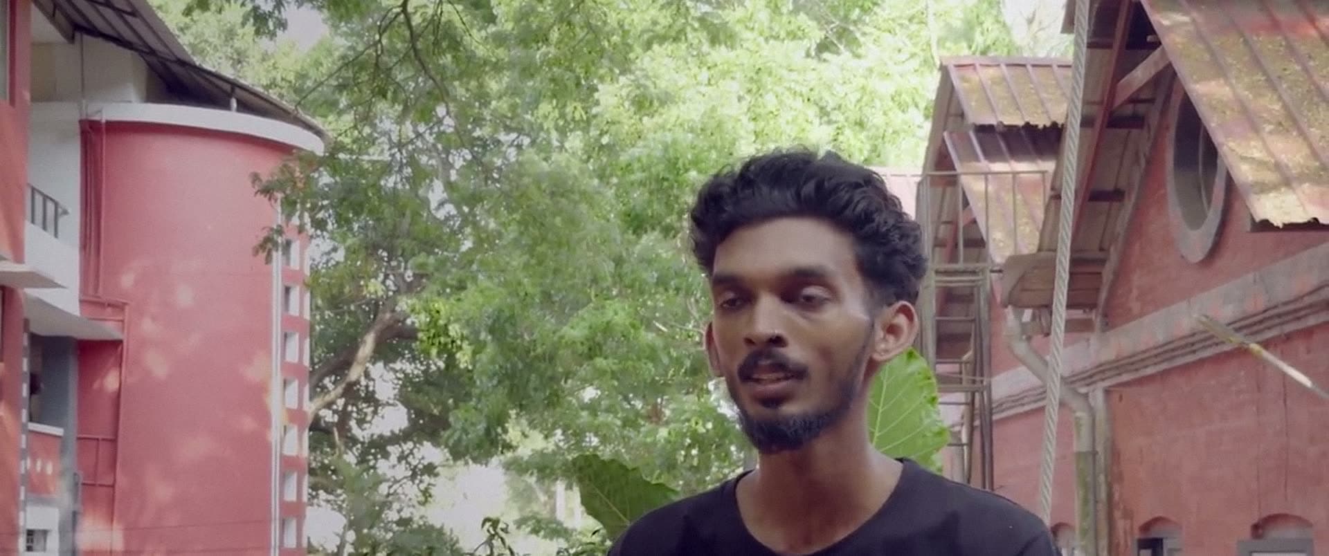 A young man speaks outdoors in front of old red-brick college buildings.