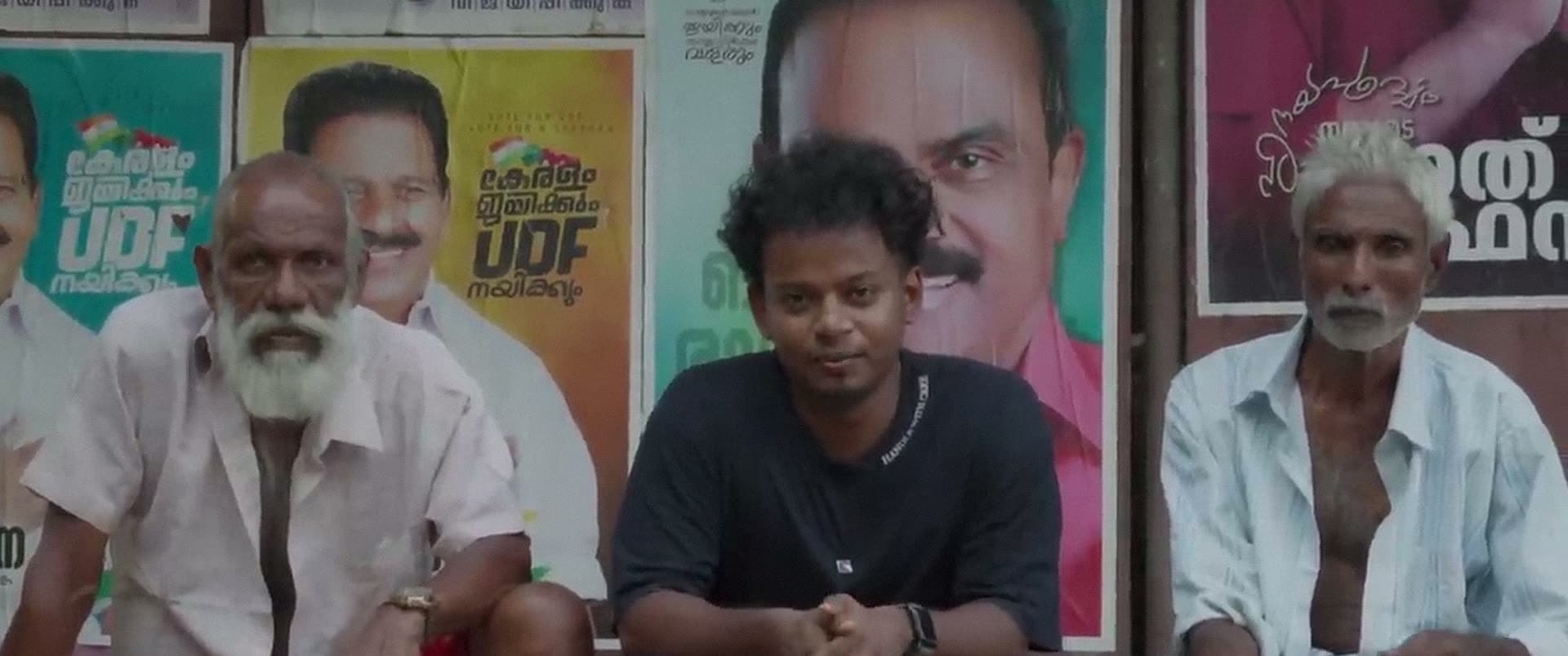 Three men sit side by side on a step, election posters covering the wall behind them.