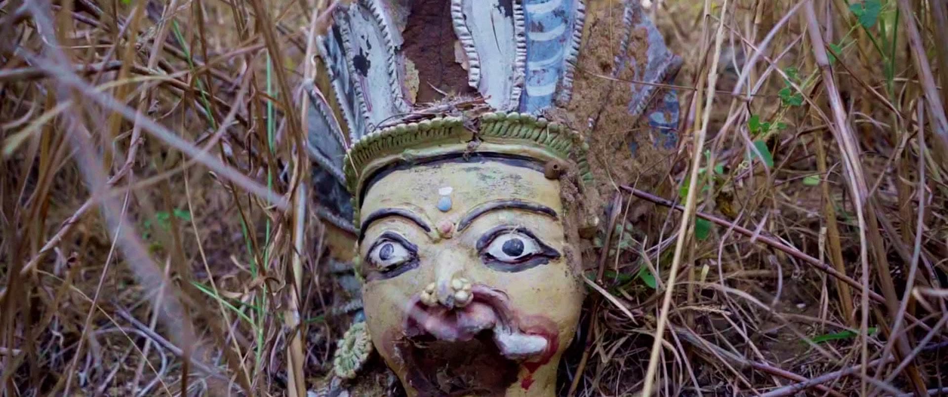A weathered painted figure with a feathered crown, surrounded by dry grass.