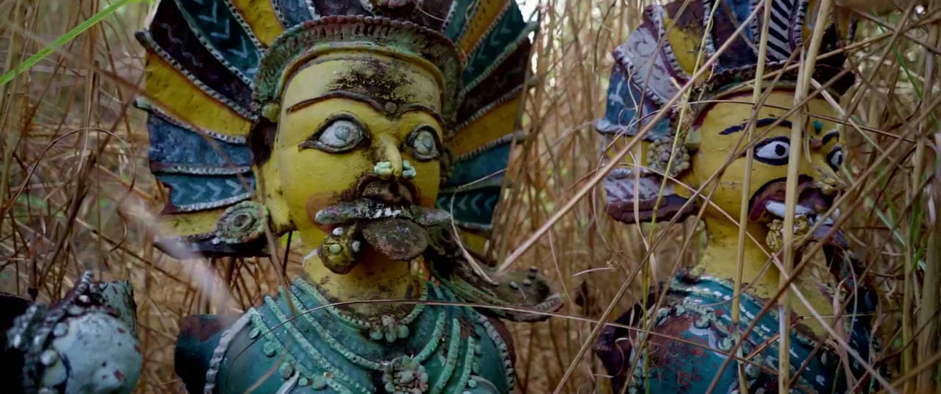 Two painted clay figures with ornate headdresses, half-hidden in tall dry grass.