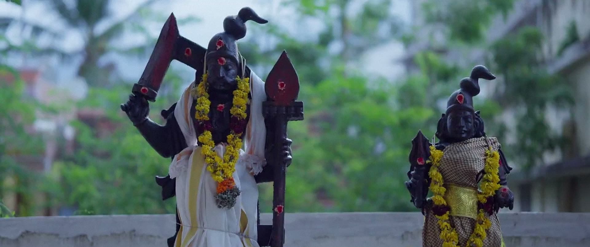 Two dark figures hung with flower garlands, standing above a wall with trees behind them.