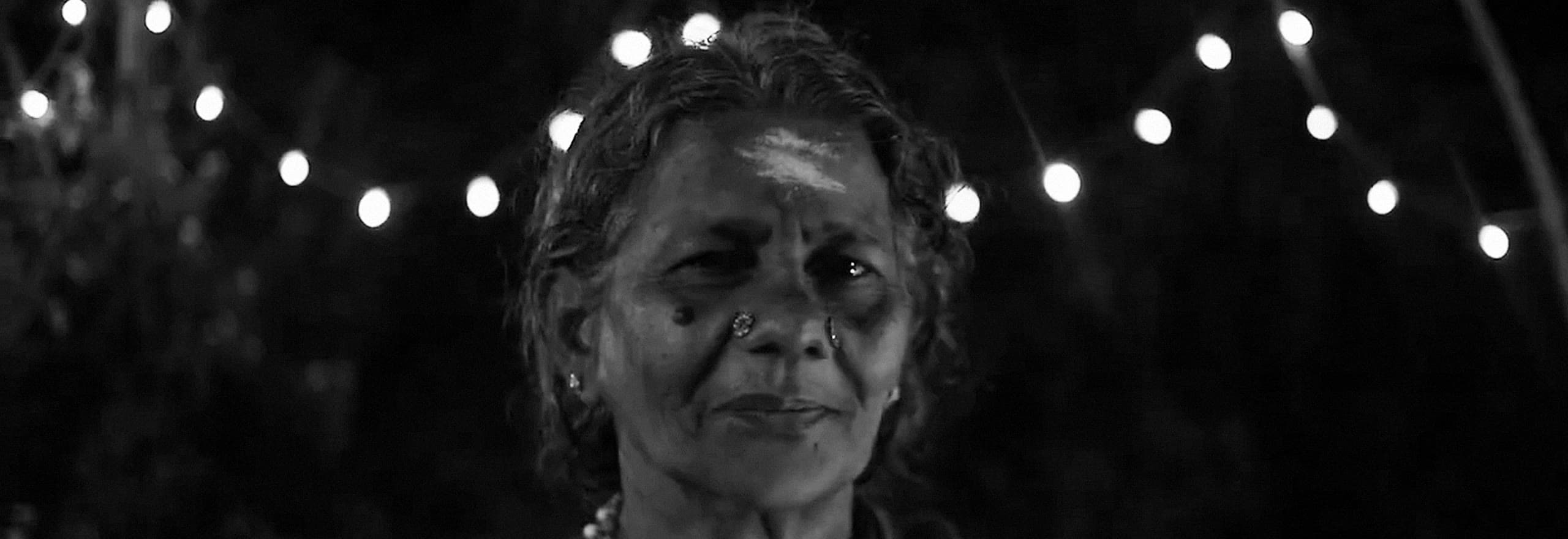 An elderly woman under strings of festival lights at night, sacred ash on her forehead.