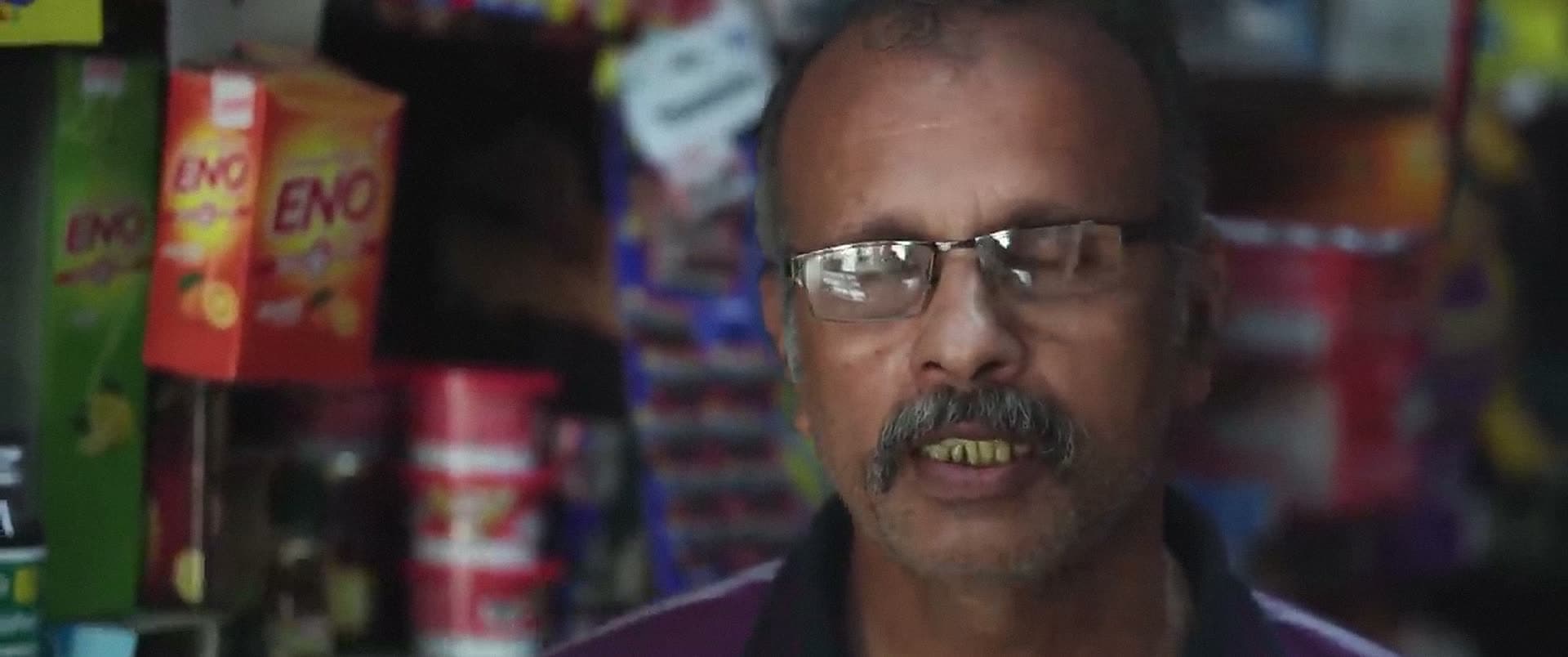 A man in glasses speaks from inside a small shop hung with packets of goods.