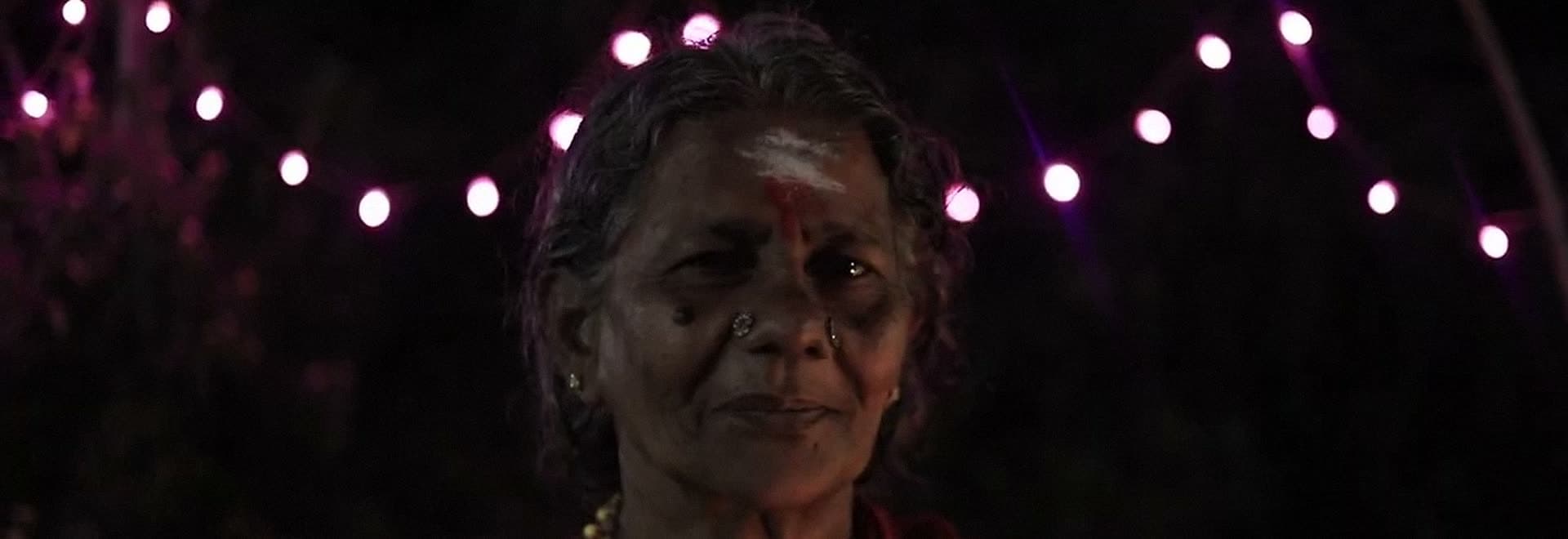 An elderly woman under strings of festival lights at night, sacred ash on her forehead.