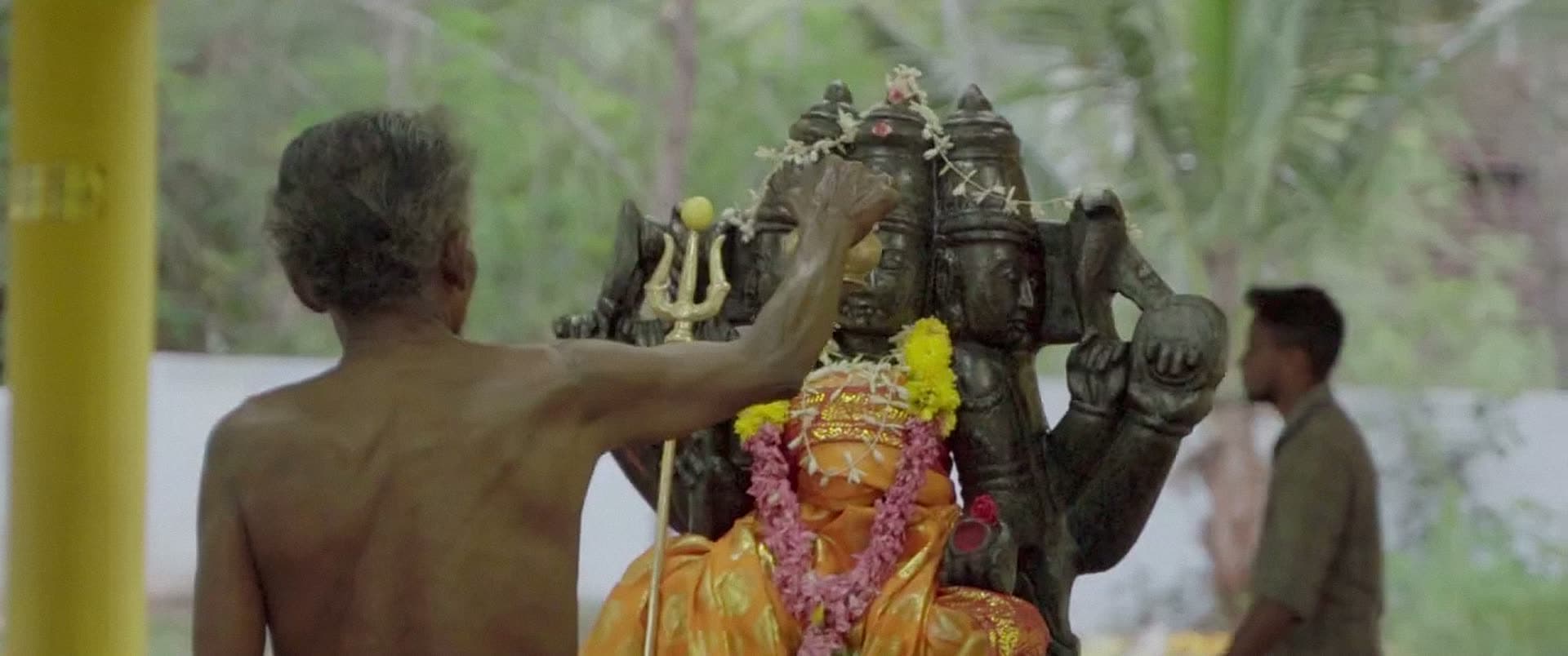 Seen from behind, a man reaches up to touch a dark stone image hung with garlands.