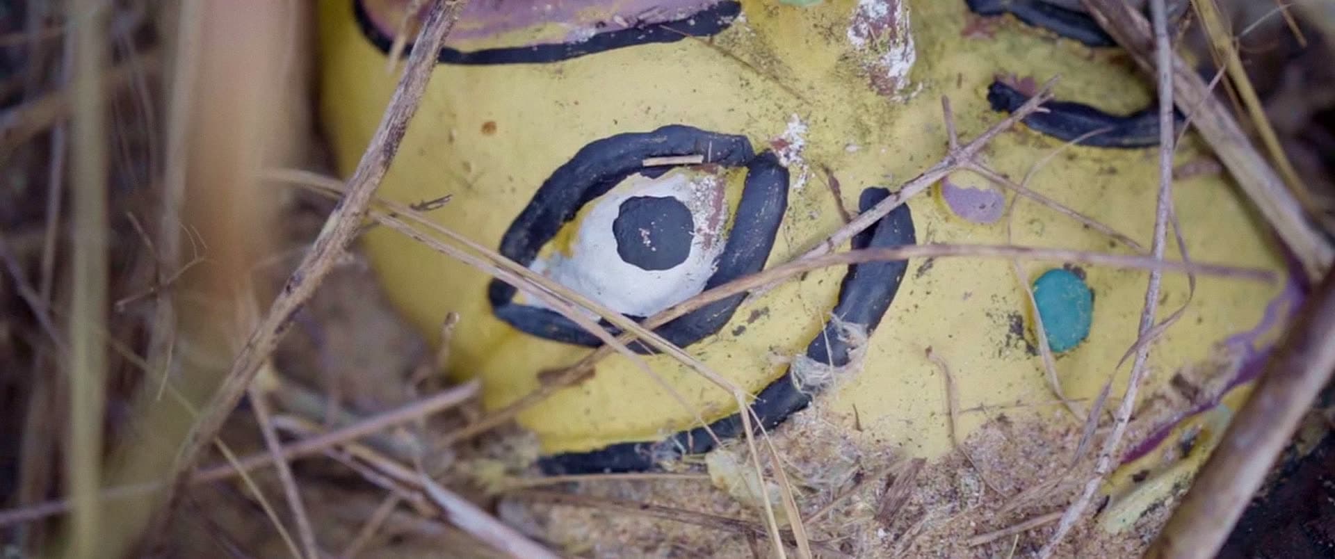 Extreme close-up of an eye painted on a weathered yellow clay figure lying in dry grass.
